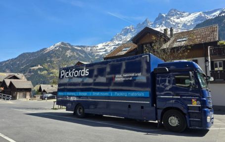 a huge blue shipping pickford truck running across the street with mountain covered in snow
